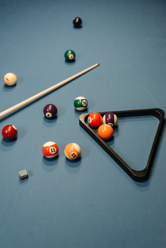 A blue pool table with several numbered balls, a cue stick, and a triangle rack scattered across the surface.